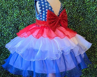 May include: A patriotic-themed dress on a mannequin. The dress features a blue bodice with white star accents, a red sequined waist, and a layered skirt in red, white, and blue. A large red bow adorns the back. The dress is displayed on a wooden stand.