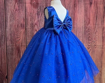 Op de afbeelding: Een koninklijk blauwe tule jurk met een satijnen lijfje en een grote strik op de rug. De jurk is versierd met kleine, glinsterende kralen.