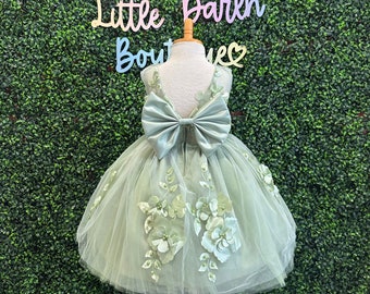 May include: A sage green tulle dress with a large satin bow on the back. The dress is decorated with 3D floral appliques.