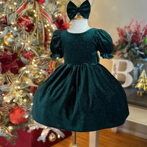 May include: A green velvet dress with a sparkly finish and short puffed sleeves. The dress is on a mannequin with a green velvet bow headband.