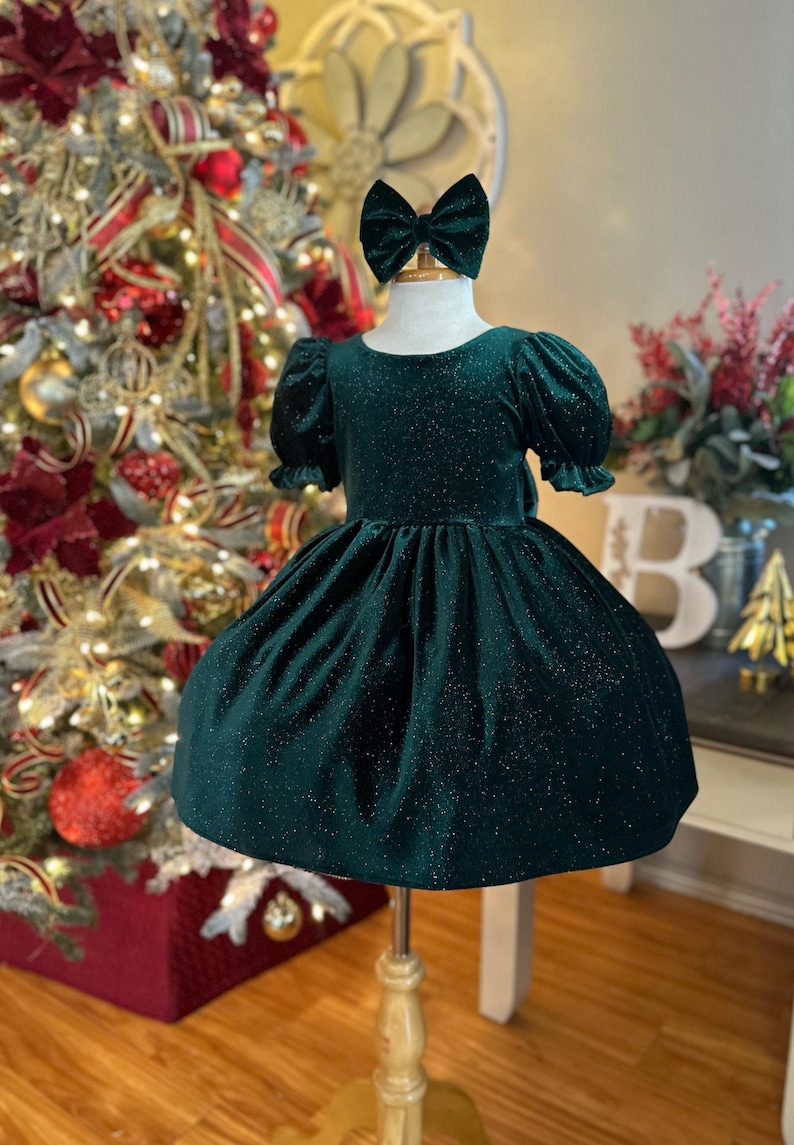 May include: A green velvet dress with a sparkly finish and short puffed sleeves. The dress is on a mannequin with a green velvet bow headband.