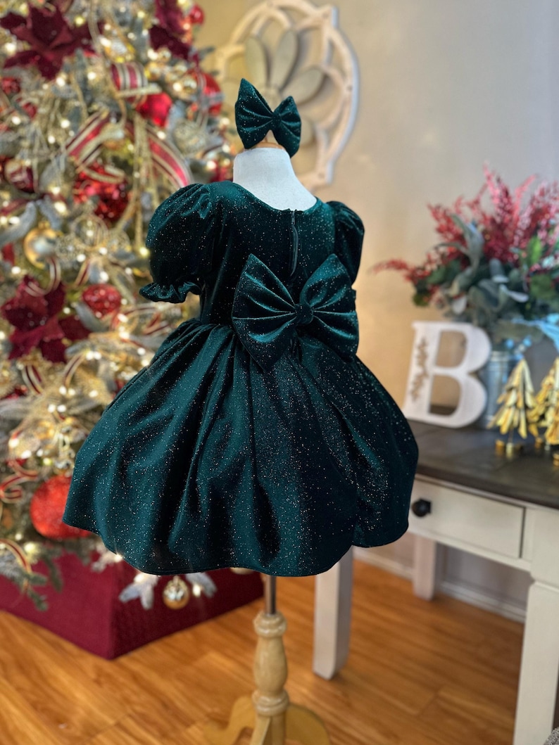 May include: A dark green velvet dress with a sparkly finish and a large bow in the back. The dress has short puffed sleeves and a full skirt.