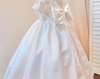 Puede incluir: Vestido blanco de satén para niña de las flores con un gran lazo en la espalda. El vestido presenta un corpiño de encaje y mangas cortas. La falda amplia crea una silueta clásica y elegante.