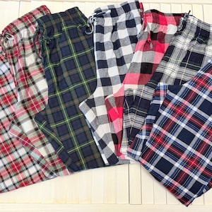 May include: Assortment of plaid pajama pants in various colors, including red, green, black, and blue. Each pair features a drawstring waist and a classic plaid pattern. The pants are folded and arranged on a white surface.