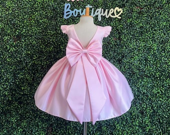 May include: A pink satin dress with a large bow on the back. The dress has a V-shaped back and short, ruffled sleeves. The dress is on a mannequin against a green backdrop.