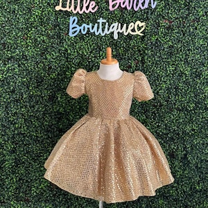 Puede incluir: Un vestido de lentejuelas doradas con cuello redondo y mangas abullonadas cortas. El vestido tiene una falda completa y es perfecto para una ocasión especial.