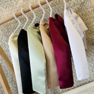 May include: Six satin fabric long-sleeved tops hanging on a wooden rack with pearl-beaded hangers. The tops are in various colors, including white, beige, green, black, and burgundy.
