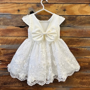 May include: A white satin and lace dress with a large bow on the back. The dress has a scalloped hem and floral embroidery.