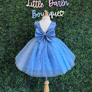 May include: A light blue tulle dress with a satin bodice and a large bow in the back. The dress is decorated with small white pearls.