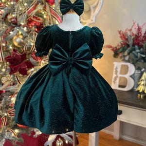May include: A green velvet dress with a sparkly finish and a large bow on the back. The dress has short puffed sleeves and a full skirt.