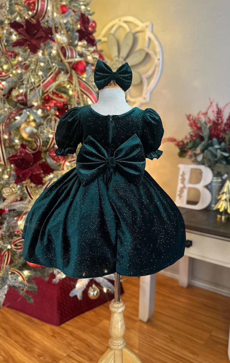 May include: A green velvet dress with a sparkly finish and a large bow on the back. The dress has short puffed sleeves and a full skirt.