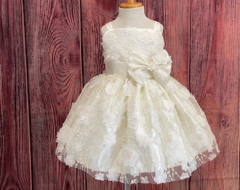 May include: Ivory-colored flower girl dress with a square neckline and spaghetti straps. The dress features a large bow at the waist and is adorned with floral appliqués. The skirt is full and layered, creating a classic look.