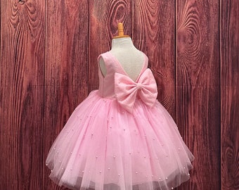 May include: A pink tulle dress with a large pink bow on the back. The dress is decorated with small white beads.