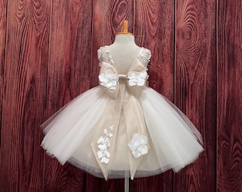 May include: A white and beige tulle dress with floral accents. The dress features a large bow on the back and decorative flowers. The dress is displayed on a mannequin, showcasing its design and details.