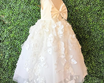 Op de afbeelding: Een witte bloemenmeisjesjurk met een grote strik op de rug. De mouwloze jurk heeft een tule rok versierd met 3D bloemen applicaties en kleine kralen. De jurk wordt op een mannequin gepresenteerd.