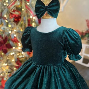May include: A green velvet dress with a sparkly texture and puffed sleeves. The dress has a round neckline and a full skirt. A green velvet bow is on top of the dress.
