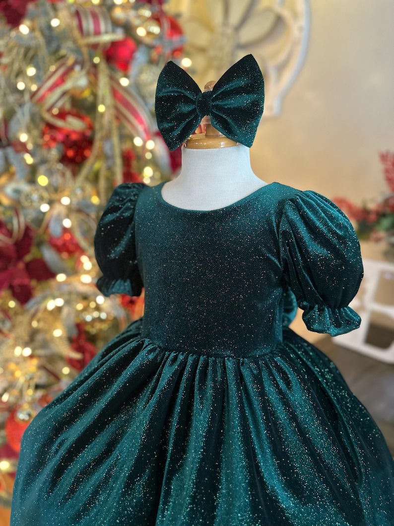 May include: A green velvet dress with a sparkly texture and puffed sleeves. The dress has a round neckline and a full skirt. A green velvet bow is on top of the dress.