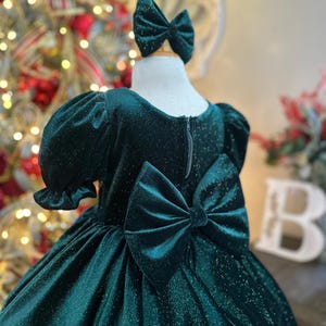 May include: A green velvet dress with a sparkly finish and a large bow in the back. The dress has short puffed sleeves and a full skirt.