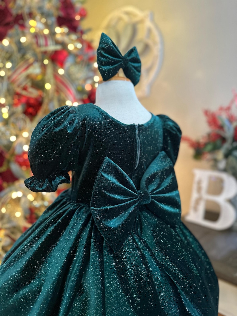 May include: A green velvet dress with a sparkly finish and a large bow in the back. The dress has short puffed sleeves and a full skirt.