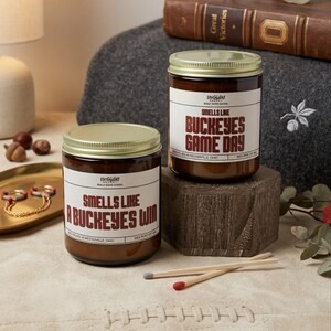 May include: Two amber glass jar candles with gold lids and white labels. One label reads "SMELLS LIKE A BUCKEYES WIN" and the other reads "SMELLS LIKE BUCKEYES GAME DAY". Two wooden matches with gray and red tips lie on the surface in front of the jars.