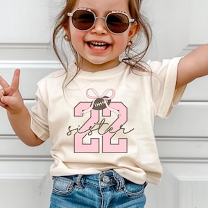 May include: A young girl wearing a light beige t-shirt with a pink number "22" and the word "sister" printed on it. The number has a brown football with a pink bow on top. She is wearing blue denim shorts and light beige sneakers. The girl is smiling and has sunglasses on.