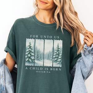 May include: Dark teal t-shirt with the text "FOR UNTO US A CHILD IS BORN ISAIAH 9:6". The graphic features a winter forest scene with snow-covered pine trees. The shirt has rolled-up sleeves.