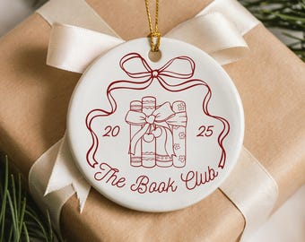 May include: A white ceramic ornament with a red design featuring three books tied with a bow, the text "The Book Club", and the numbers 20 and 25. The ornament hangs from a gold string and rests on a wrapped gift.
