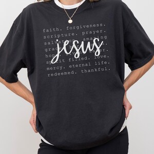 May include: Black t-shirt with the word "Jesus" in a cursive font, surrounded by words like "faith," "forgiveness," and "love." The shirt has a white crew neck and is worn with black shorts.