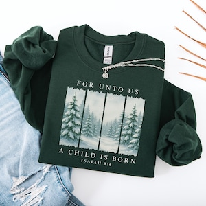 May include: A dark green sweatshirt with the text "FOR UNTO US A CHILD IS BORN ISAIAH 9:6" and a winter forest scene. A silver necklace with a pendant is on the sweatshirt. The sweatshirt is next to a pair of blue jeans.