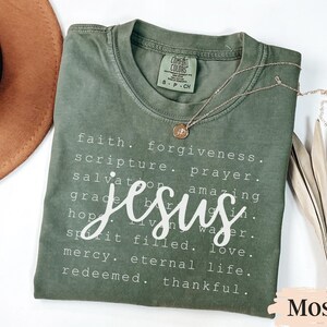 May include: A moss green t-shirt with the word "Jesus" in a white script font. The shirt also features a list of words like "faith," "forgiveness," and "love." A gold necklace with a pendant rests on the shirt.
