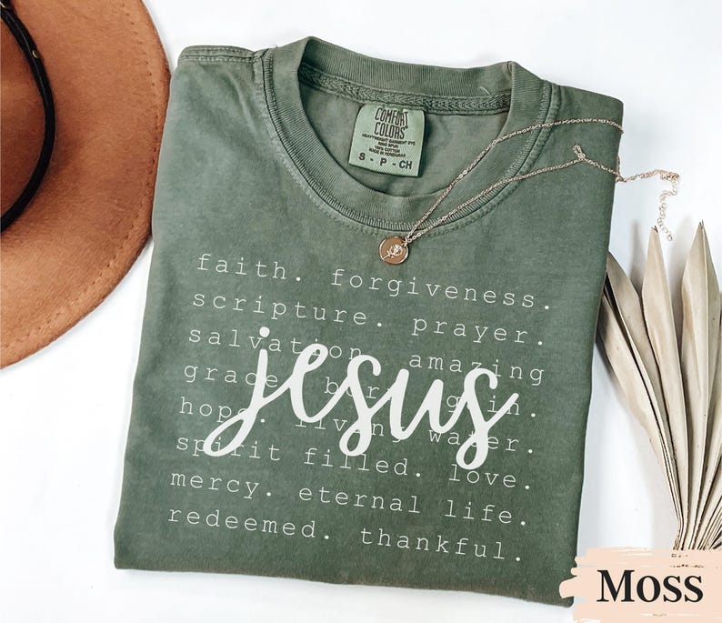 May include: A moss green t-shirt with the word "Jesus" in a white script font. The shirt also features a list of words like "faith," "forgiveness," and "love." A gold necklace with a pendant rests on the shirt.