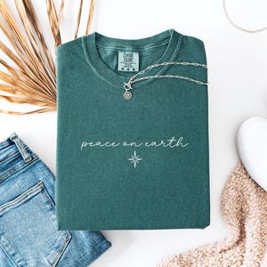 May include: A teal Comfort Colors t-shirt with the words "peace on earth" and a star graphic. A silver necklace with a circular pendant rests on the shirt. The shirt is paired with blue jeans, white sneakers, and a beige scarf.