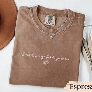 May include: Espresso-colored t-shirt with the phrase "falling for jesus" in white script, along with a maple leaf graphic. A silver necklace with a pendant rests on the shirt. The shirt has a crew neck and a Comfort Colors tag.