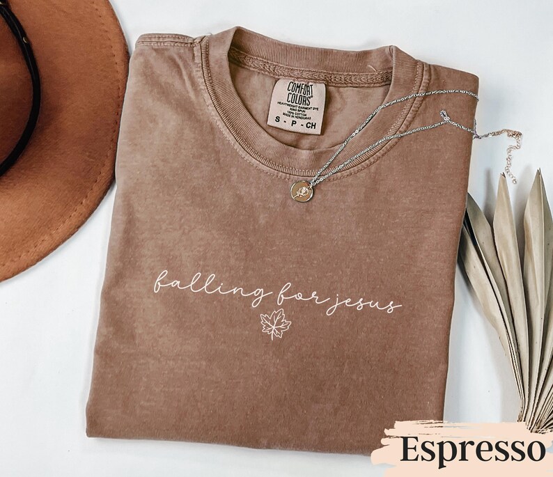 May include: Espresso-colored t-shirt with the phrase "falling for jesus" in white script, along with a maple leaf graphic. A silver necklace with a pendant rests on the shirt. The shirt has a crew neck and a Comfort Colors tag.