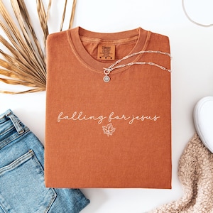 May include: Rust-colored t-shirt with the phrase "falling for jesus" in white script and a leaf graphic. A silver necklace and blue jeans are next to the shirt. The shirt is folded and has a tag that says "Comfort Colors".