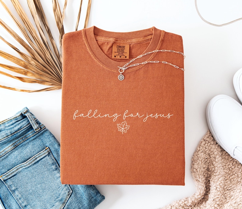 May include: Rust-colored t-shirt with the phrase "falling for jesus" in white script and a leaf graphic. A silver necklace and blue jeans are next to the shirt. The shirt is folded and has a tag that says "Comfort Colors".
