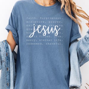 May include: A blue t-shirt with the word "Jesus" in a cursive font, surrounded by words like "faith," "forgiveness," and "eternal life." The shirt has rolled-up sleeves and the text "Blue Jean" at the bottom.