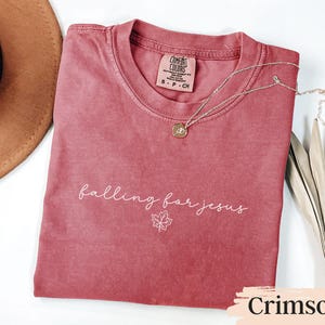 May include: Crimson-colored t-shirt with the phrase "falling for jesus" in white script and a maple leaf graphic. A gold necklace with a pendant rests on the shirt. The shirt is folded, with a hat and dried leaves in the background.