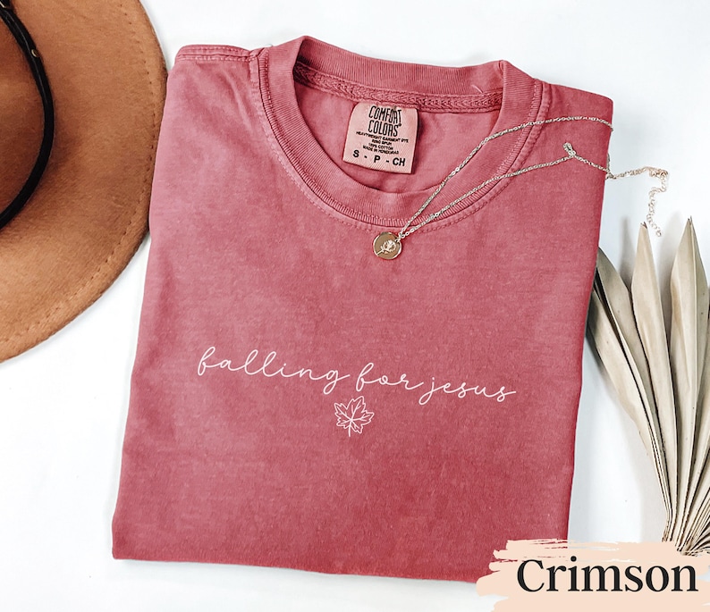 May include: Crimson-colored t-shirt with the phrase "falling for jesus" in white script and a maple leaf graphic. A gold necklace with a pendant rests on the shirt. The shirt is folded, with a hat and dried leaves in the background.