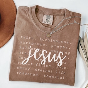 May include: A brown Comfort Colors t-shirt with the word "Jesus" in white script. The shirt features a list of words including "faith", "forgiveness", and "love". A silver necklace with a small pendant is draped on the shirt.