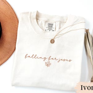 May include: Ivory-colored t-shirt with the phrase "falling for jesus" in a brown script font, accompanied by a maple leaf graphic. A gold necklace with a pendant rests on the shirt. The shirt is labeled "Comfort Colors".