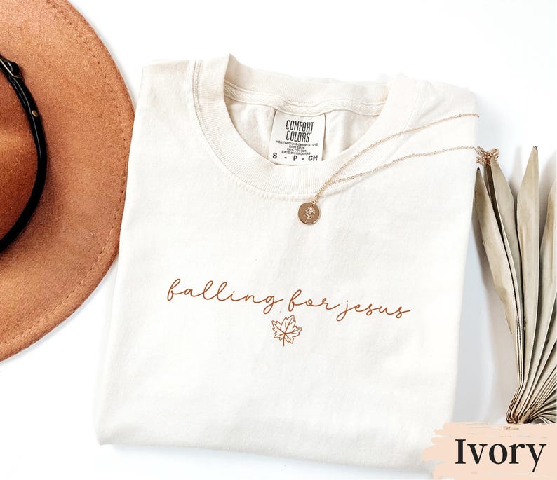 May include: Ivory-colored t-shirt with the phrase "falling for jesus" in a brown script font, accompanied by a maple leaf graphic. A gold necklace with a pendant rests on the shirt. The shirt is labeled "Comfort Colors".