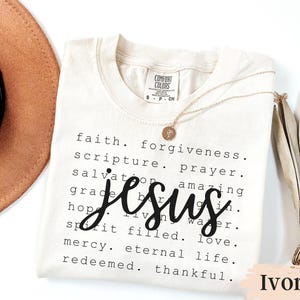 May include: Ivory-colored t-shirt with the word "Jesus" in a large, cursive font. The shirt also features a list of related words in a smaller font. A gold necklace with a pendant rests on the shirt.