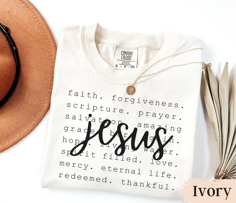 May include: Ivory-colored t-shirt with the word "Jesus" in a large, cursive font. The shirt also features a list of related words in a smaller font. A gold necklace with a pendant rests on the shirt.