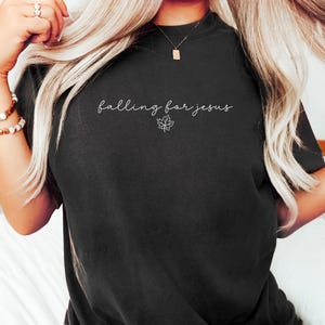 May include: Black t-shirt with the phrase "falling for jesus" in cursive script and a small maple leaf graphic. The word "Black" is printed at the bottom of the shirt. A casual, comfortable style.