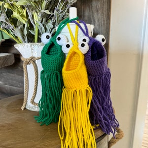 May include: Three crocheted hanging ornaments with fringe, each in a different color: green, yellow, and purple. Each ornament has two large black eyes.