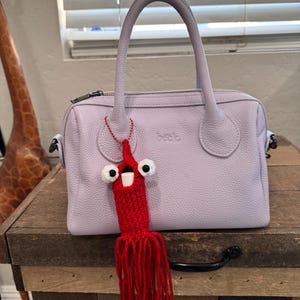 May include: A lavender leather handbag with a red crocheted octopus keychain. The keychain has white googly eyes and a red tassel.
