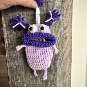 May include: A handmade crochet monster in shades of purple and lavender. The whimsical creature has large eyes, a wide mouth, and tufts of hair. It is suspended by a white string and held by a hand against a wooden background.