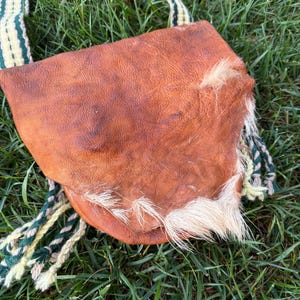 May include: A brown leather shoulder bag with a flap closure and a woven strap. The bag features a patch of white fur on the side. The strap is green and white. The bag is lying on green grass.