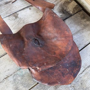 May include: A brown leather satchel with a flap closure and a long, adjustable strap. The satchel has a worn, distressed appearance, with visible stitching and a brass buckle. The bag is designed to be worn over the shoulder.